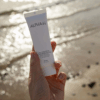 Hand holding Alpha-H Daily Essential Moisturiser SPF 50 against a beach-inspired background, highlighting sun protection and hydration.