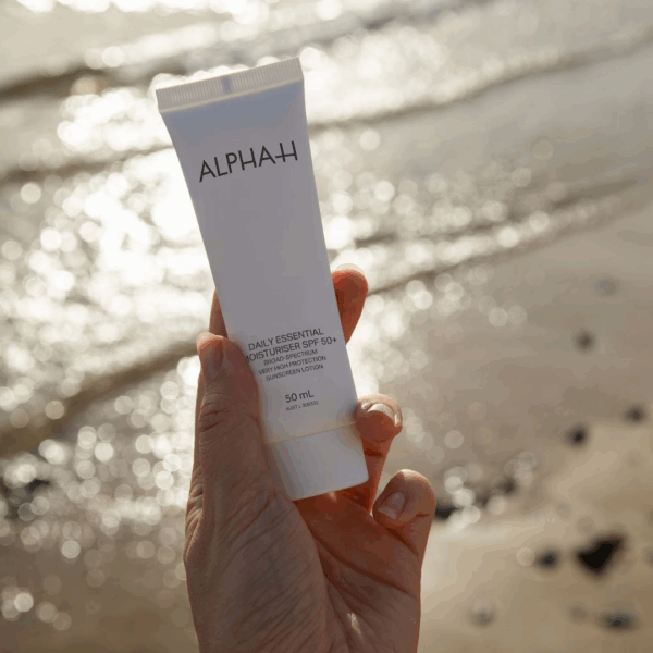 Hand holding Alpha-H Daily Essential Moisturiser SPF 50 against a beach-inspired background, highlighting sun protection and hydration.