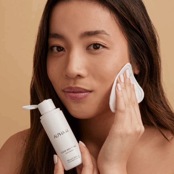 Woman applying Alpha H Clear Skin Tonic 100ml with a cotton pad on her face, helping to exfoliate and refine skin texture.